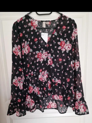 NY Blommig svart blus från H&M med etikety - Snygg svart blus från H&M med romantiskt blommönster i rosa och rött. Blusen är i ett tunt, transparent material och har volangdetaljer vid ärmsluten. Perfekt för dig som gillar en feminin och trendig stil.