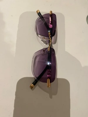 Lila solglasögon med gulddetaljer - Snygga solglasögon med lila, rektangulära glas och guldfärgade detaljer på skalmarna. Skalmarna är svarta med guldfärgade ändar och har en unik dekoration nära gångjärnet. Perfekt accessoar för dig som vill sticka ut.