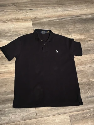 Svart piké från Polo Ralph Lauren - Klassisk svart pikétröja från Polo Ralph Lauren i modellen Custom Slim Fit. Tröjan har korta ärmar, krage med knappar och den ikoniska broderade loggan på bröstet. Perfekt till jeans eller shorts för en clean och stilren look.