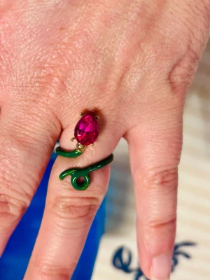 Grön ring med fucsia droppe - Unik ring i form av en grön snirklig slinga med en fucsia droppformad sten framtill. Ringen har guldfärgade detaljer runt stenen och en modern, lekfull design som sticker ut. Perfekt för dig som vill ha något färgglatt och annorlunda.