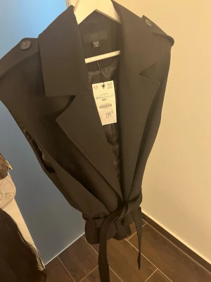 Svart ärmlös kavaj från Zara - Stilren svart ärmlös kavaj från Zara med breda slag, knytskärp i midjan och axeldekorationer med knappar. Perfekt för en trendig look med elegant känsla. Materialet är slätt och har en lätt glans.
