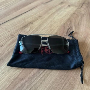 Katsuma solglasögon med röda detaljer - Snygga solglasögon från Vasuma med rektangulära, mörka glas och metallbågar i silver. Skalmarna har röda detaljer och logga på sidan. Perfekt accessoar för att lyfta din stil i sommar.