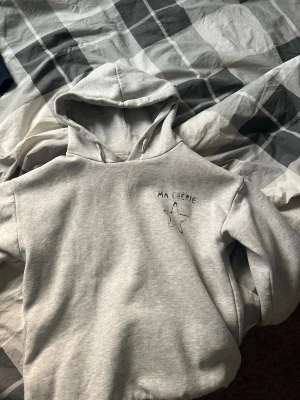 Grå hoodie med MA CHÉRIE brodyr - Säljer en ljusgrå hoodie med broderad text 'MA CHÉRIE' och en stjärna på bröstet. Hoodien har klassisk huva med dragsko och är tillverkad i mjukt material, perfekt för chill dagar. Enkel och stilren design som passar till jeans eller joggers.