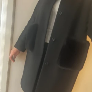 Custommade  - Väldigt fin svart kappa från Custommade med pälsdetaljer på fickor och rygg. Kappan har dekorativa blommiga knappar och rak passform. Superfin kvalite!