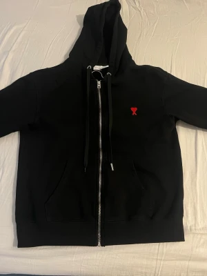 Svart zip hoodie från Ami Paris - Säljer en svart hoodie med dragkedja från Ami Paris. Hoodien har en huva med snören, känguruficka och det klassiska röda Ami-hjärtat broderat på bröstet. Tillverkad i mjuk bomull för skön känsla och stilren look. Ena fickan är sönder! Se bild annars fint skick