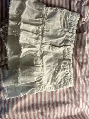 Vit volangkjol med råa kanter - Supersöt vit kortkjol med volanger och råa, fransiga kanter. Kjolen har fickor fram och bak samt knapp och dragkedja i midjan. Perfekt för dig som gillar en trendig och avslappnad stil.