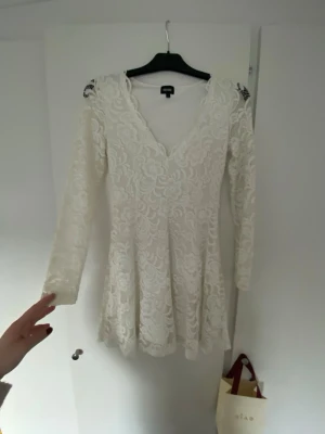 Vit spetsklänning med v-ringning - Säljer en vit kort spetsklänning med v-ringning och långa ärmar. Klänningen har ett blommigt spetsmönster och sitter snyggt i midjan med lätt utsvängd kjol. Perfekt för studenten