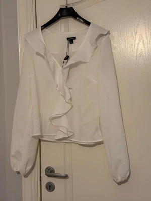 Vit volangblus från Boohoo - Stilren vit blus från Boohoo med prislapp på, med långa ärmar och volangdetalj framtill. Blusen har en bred krage.  Perfekt för dig som gillar en fin och trendig look.