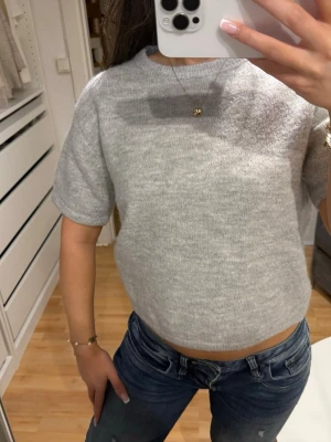 Grå kortärmad stickad topp - Säljer en enkel och stilren grå stickad topp med korta ärmar och rund hals. Modellen är något croppad och har en mjuk, bekväm känsla. Perfekt att matcha med jeans eller kjol för en avslappnad look. Aldrig använt 
