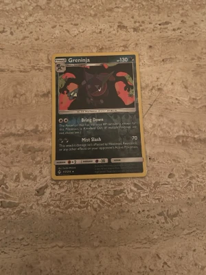 Pokémonkort Greninja - Greninja Pokémonkort med svart, röd och grön illustration. Kortet har 130 HP och attacker som Bring Down och Mist Slash. Baksidan har klassiskt blått Pokémon-motiv med pokéboll. Perfekt för samlare eller Pokémonfans.