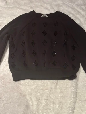 Svart sweatshirt med paljettdetaljer - Svart sweatshirt med glittrande paljettdetaljer framtill. Tröjan har rund halsringning, långa ärmar och ribbade muddar vid ärmslut och nederkant. Perfekt för dig som vill ha en enkel men ändå snygg look. Materialet känns mjukt och bekvämt.