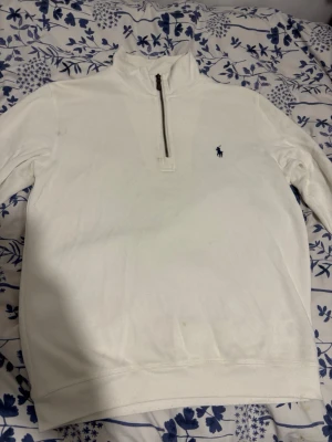 Vit halvzip tröja från Ralph Lauren - Klassisk vit tröja med halv dragkedja från Ralph Lauren. Tröjan har lång ärm, hög krage och den ikoniska blå loggan broderad på bröstet. Perfekt för en clean och stilren look, lätt att matcha med jeans eller chinos.      OBS!! Den har små fläckar som går säkert att få bort på något sätt ni kan se dem på bilden.