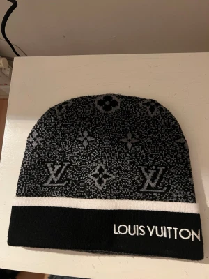 Svart och grå mössa - Snygg svart och grå mössa med klassiskt monogrammönster och vit logotyp på uppvikt kant. Mössan har en kontrasterande vit rand och är stickad i mjukt material, perfekt för att hålla värmen och ge en exklusiv touch till din outfit.