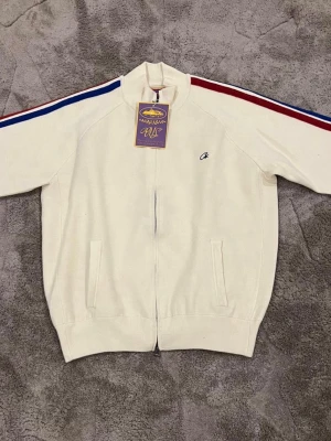 Vit zip hoodie med ränder - Vit hoodie med dragkedja. Har blå och röda ränder längs ärmarna och två fickor framtill. Liten broderad logga på bröstet och ribbade muddar. Perfekt för en sportig och clean look.