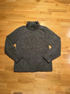 Mörkgrön vindjacka med hög krage - Använd endast ett fåtal gånger. Stilren mörkgrön vindjacka med hög krage och tryckknappar framtill. Två bröstfickor med lock och knappar samt justerbara ärmslut. Jackan har en rak passform och är tillverkad i ett lätt, vindtätt material som passar perfekt för blåsiga dagar. 