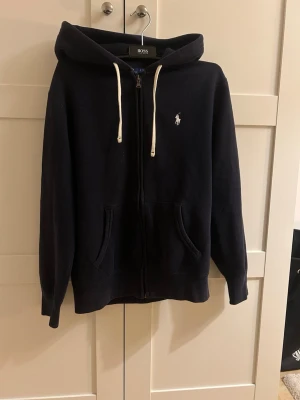 Mörkblå hoodie med dragkedja från ralph lauren - Snygg mörkblå hoodie från Ralph lauren med dragkedja framtill och vit logga på bröstet. Har huva med vita snören och två fickor fram. Perfekt för en chill och stilren look. Mjuk bomullskänsla och klassisk passform.