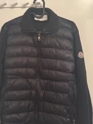 Svart dunjacka från Moncler - Snygg svart dunjacka från Moncler med quiltad design och dragkedja framtill. Jackan har ribbad krage och ärmar samt Moncler-logga på ena ärmen. Perfekt för kalla vinterdagar och riktigt stilren look.