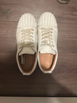Vita sneakers med nitar från Louboutin - Unika vita sneakers från Christian Louboutin med coola nitar över hela tån. Skorna är i läder med klassisk låg modell och har vita snören. Insidan är beige och sulan är vit. Perfekta för dig som vill sticka ut med din stil. Låda tillkommer