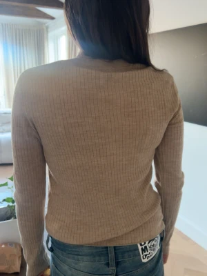 Beige ribbstickad tröja - Säljer en beige ribbstickad tröja med rund hals och långa ärmar. Tröjan är figurnära och har ett klassiskt, stilrent snitt som passar perfekt till jeans. Materialet är mjukt och stretchigt, vilket gör den bekväm att bära. Perfekt för dig som gillar enkel och snygg design.