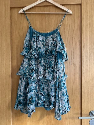 Blommig volangklänning från Zara - Supersöt kort blommig klänning från Zara med tunna axelband och flera lager volanger. Klänningen har en mix av blå, beige och svarta toner med inslag av guldiga detaljer. Perfekt för sommarens alla tillfällen!