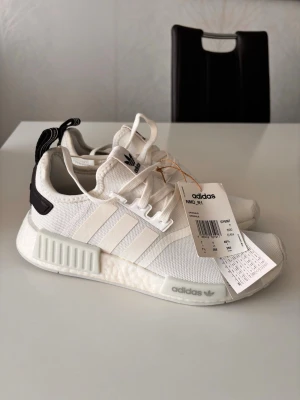Adidas NMD_R1 vita sneakers - Fräscha Adidas NMD_R1 sneakers i vitt med svarta detaljer på hälen och klassiska tre ränder på sidan. Ovandelen är i mesh och textil för en luftig känsla, och sulan har Boost-dämpning för extra komfort. Snygg och modern siluett som passar till det mesta. Helt nya, aldrig använda då det var fel storlek.