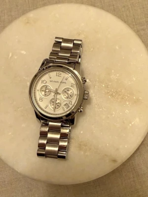 Silverfärgad klocka Michael Kors - Stilren klocka från Michael Kors i silverfärgat metallarmband och rund urtavla. Klockan har flera små urtavlor, tydliga siffror och datumvisning. Perfekt accessoar för dig som gillar klassisk och modern design. 