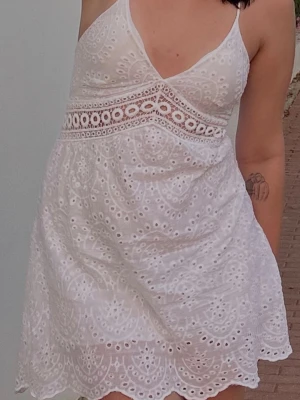 Vit kort klänning med spetsdetaljer - Söt vit kort klänning med broderade spetsdetaljer och dekorativa hålmönster. Klänningen har smala axelband med knytning och v-ringad urringning. Perfekt för sommarens varma dagar och har en feminin, romantisk stil. Köpt i Italien, ingen storlek men passar S