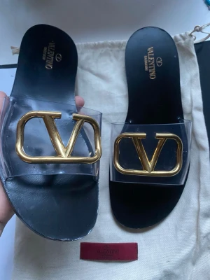 Valentino sandaler med guldlogo - Snygga svarta sandaler från Valentino med bred transparent rem och stor guldfärgad V-logga framtill. Sulan är i svart läder och remmen i plast ger en modern touch. Perfekta för dig som vill ha en exklusiv och trendig look.