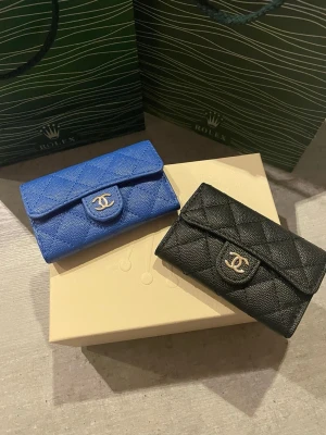 quiltade plånböcker blå & svart - Två stilrena plånböcker i quiltat läder, en i klarblått och en i klassiskt svart. Båda har det ikoniska CC-spännet i silver framtill och praktisk knappstängning. Perfekta accessoarer för dig som gillar lyxig och tidlös design.