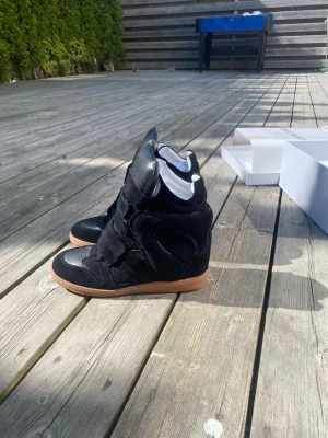 Isabel Marant svarta wedge sneakers - Svarta sneakers från Isabel Marant med högt skaft och inbyggd kilklack. Skorna har en mix av mocka och läder, breda kardborreband och en klassisk gummisula i beige. Perfekta för dig som vill ha en edgy och trendig look.