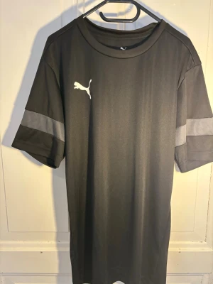 Svart tränings t-shirt från Puma - Snygg svart t-shirt från Puma med klassisk logga på bröstet och gråa detaljer på ärmarna. T-shirten har rund halsringning och är tillverkad i ett mjukt, sportigt material som passar perfekt till en aktiv stil. (Inte använd bara testad)