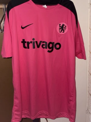 Chelsea tränings t-shirt rosa Nike XL - Chelsea tränings t-shirt i rosa med svarta detaljer från Nike. Trivago-logga och Chelsea FC-emblem på bröstet. Tillverkad i lätt och ventilerande material, perfekt för träning eller match. Klassisk rund hals och korta ärmar.