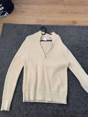 Beige stickad tröja med halv dragkedja - Säljer en beige stickad tröja med halv dragkedja och ståkrage. Tröjan har ett diskret strukturmönster, långa ärmar och ribbade muddar vid ärmslut och nederkant. Perfekt för lager-på-lager och enkel att matcha.