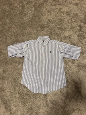 Randig skjorta från Ralph Lauren - Klassisk skjorta från Ralph Lauren med blå och vita ränder, korta ärmar och button-down krage. Skjortan har den ikoniska broderade loggan på bröstet och stängs med vita knappar. Tillverkad i mjuk bomull för en bekväm känsla. Passar som 158-164