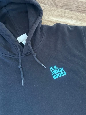 Svart hoodie N.Y. Neighbours - Svart hoodie från N.Y. Neighbours med turkos text på bröstet och en stor grafisk ryggprint i turkost och svart. Klassisk huva med snören och mjukt material, perfekt för en avslappnad streetwear-look. Priset går att diskutera!!