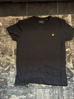 Svart t-shirt från Lyle & Scott - Svart t-shirt från Lyle & Scott med klassisk passform och rund hals. T-shirten har en gul broderad logga på bröstet och är tillverkad i mjuk bomull. Perfekt för en stilren och enkel look.