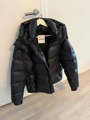 Svart pufferjacka från J.Lindeberg - Stilren svart pufferjacka från J.Lindeberg med hög krage och avtagbar huva. Jackan har breda kanaler, dragkedja framtill och tryckknappar. Perfekt för kalla dagar och ger en modern look. Materialet är glansigt och vattenavvisande.