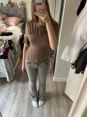 Brun korsettinspirerad topp - Snygg brun topp med korsettinspirerade sömmar och korta ärmar. Toppen har en rund halsringning och lätt rynkade detaljer över bysten som ger en cool form. Perfekt att styla med jeans eller kjol för en trendig look.