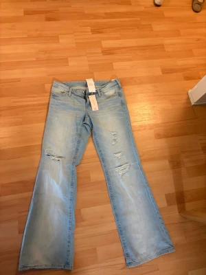 Ljusblå bootcut jeans med slitningar - Säljer ett par ljusblå bootcut jeans med trendiga slitningar på framsidan. Jeansen har klassisk femficksmodell och är tillverkade i denim. Perfekta för en avslappnad och cool stil.