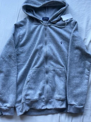 Helt ny  Polo Ralph Lauren - Helt ny grå hoodie från Polo Ralph Lauren med dragkedja, huva och kängurufickor. Storlek S men passar även XS. Tröjan har inga täcken på användning då jag aldrig använt den. Pris kan dirkuteras vid snabb affär.