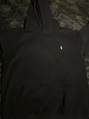 Svart hoodie från Polo Ralph Lauren - Svart hoodie från Polo Ralph Lauren med klassisk huva och känguruficka. Liten vit broderad logga på bröstet. Perfekt för en chill och stilren look. Mjuk bomullskänsla och enkel att matcha med det mesta.