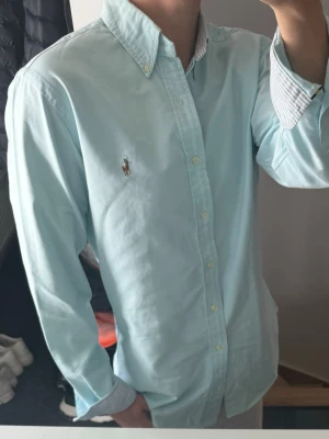 Ljusblå skjorta från Ralph Lauren - Klassisk ljusblå skjorta från Ralph Lauren med button-down krage och broderad logga på bröstet. Skjortan har långa ärmar, vita knappar och är tillverkad i mjuk bomull. Perfekt för en clean och stilren look.