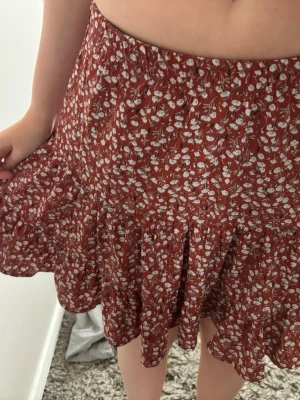 Rödblommig volangkjol - Söt röd kjol med vitt blommönster och volangkant. Kjolen har en avslappnad passform med resår i midjan och är tillverkad i ett lätt och luftigt tyg, perfekt för varma dagar.