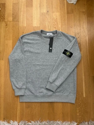 Grå sweatshirt från Stone Island - Snygg grå sweatshirt från Stone Island med klassisk rund hals och ribbade muddar. Tröjan har den ikoniska Stone Island-loggan på vänster ärm och är tillverkad i mjukt bomullsmaterial. Perfekt för en clean och stilren look.