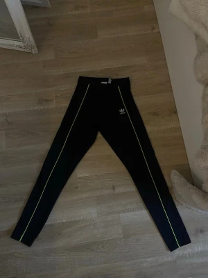 Svarta leggings från Adidas XS - Svarta leggings från Adidas med smala neongula ränder längs benen och klassisk Adidas-logga på höften. Tight passform och stretchigt material som sitter snyggt. Perfekta för en sportig och trendig look.