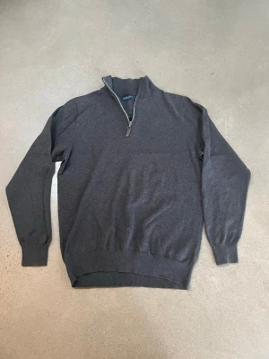 Mörkgrå half zip-tröja från Morris - Mörkgrå stickad tröja från Morris med half zip-dragkedja och hög krage. Tröjan har en klassisk passform, ribbade muddar och är perfekt för lager-på-lager-stil. Enkel och stilren design som funkar till det mesta.