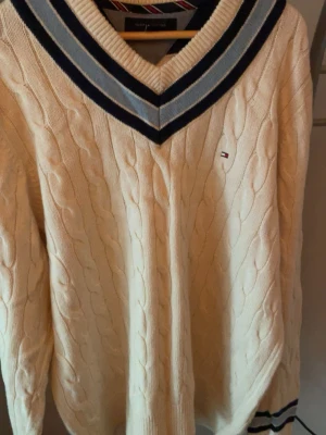 Vit kabelstickad tröja Tommy Hilfiger - Kabelstickad vit tröja från Tommy Hilfiger med V-ringning och blåa ränder runt halsen. Liten logga på bröstet och ribbade muddar. Klassisk preppy stil, perfekt för lager-på-lager och snygg till jeans.