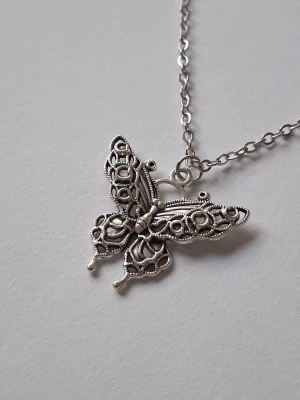 Silvrigt halsband med fjärilshänge - halsband med ett detaljerat fjärilshänge. Hänget har ett genombrutet, ornamenterat mönster som ger en vintagekänsla. Kedjan är tunn och stilren, perfekt för dig som gillar unika accessoarer med naturinspirerad design. Materialet rostfritt stål. 