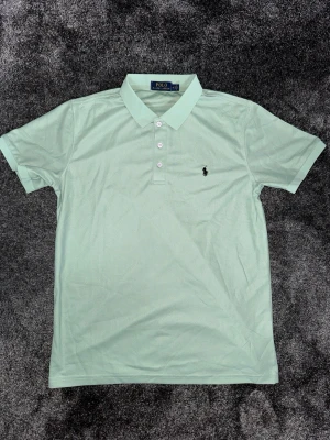 Ralph Lauren Piké strl M - Fräsch mintgrön pikétröja från Polo Ralph Lauren med klassisk krage och tre knappar framtill. Tröjan har korta ärmar och den ikoniska broderade loggan på bröstet. Perfekt för en stilren och avslappnad look. Storlek M, endast använd ett fåtal gånger.