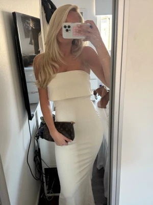 Vit axelbandslös långklänning - Säljer en elegant beige långklänning med axelbandslös design och figurnära passform. Klänningen har en enkel och stilren look med en volang upptill. Perfekt för balen men hittade en annan jag ville ha💞💞den här prislapp o allt kvar